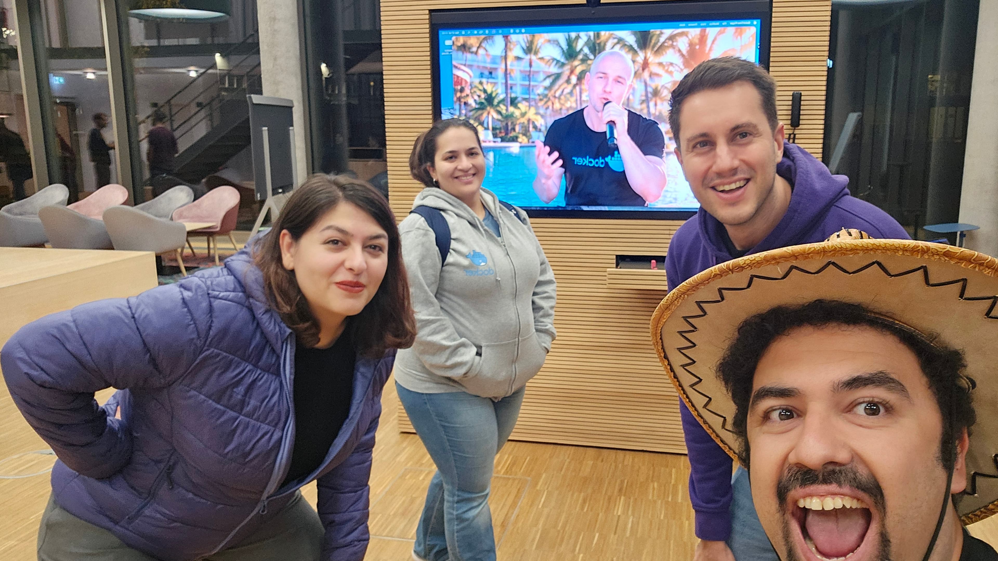 Four attendees pose for a selfie in front of a screen showing a Docker video; the person in front wears a straw hat.