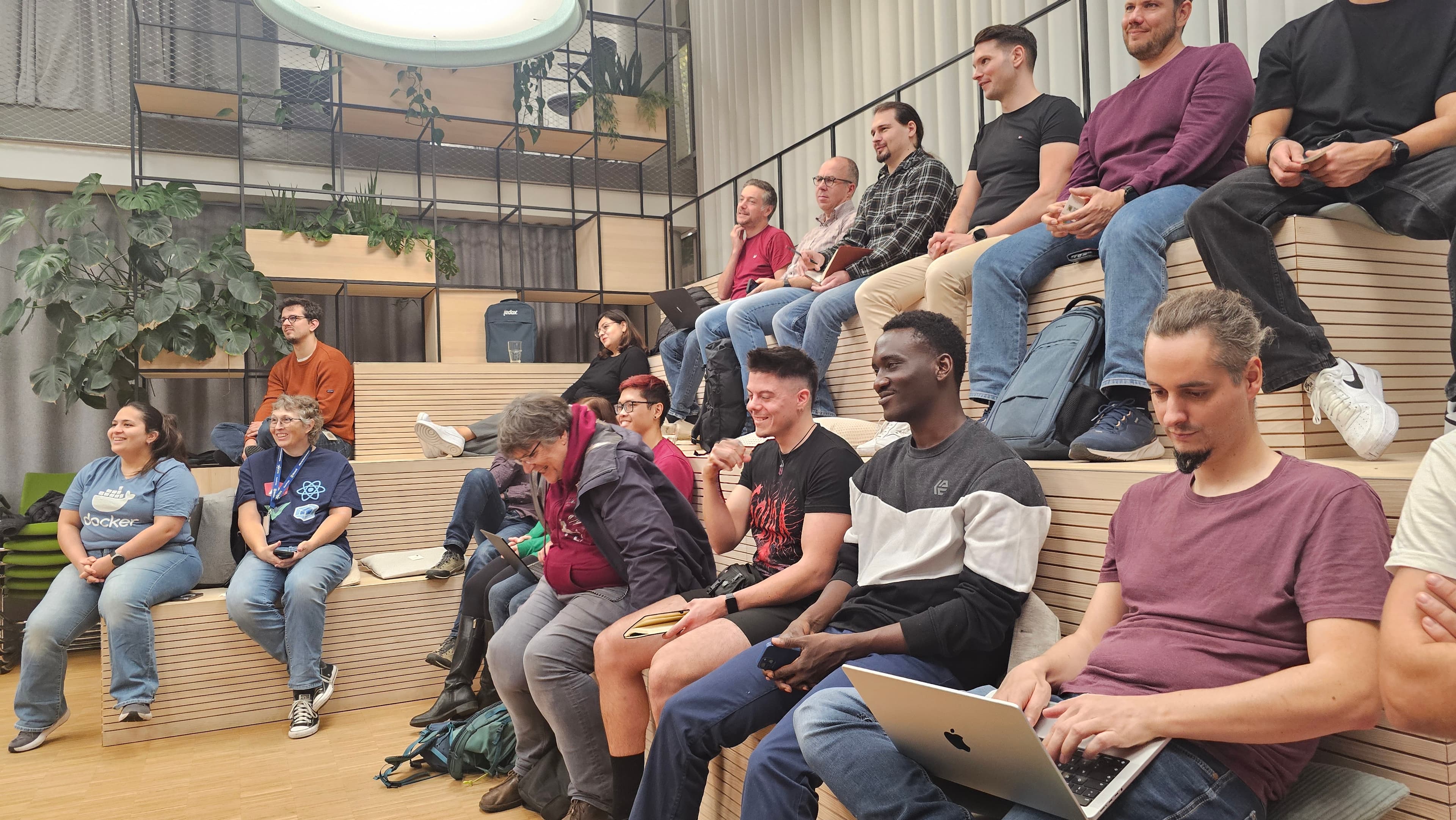 Attendees smile and laugh on tiered wooden seating, with a laptop open in the foreground.
