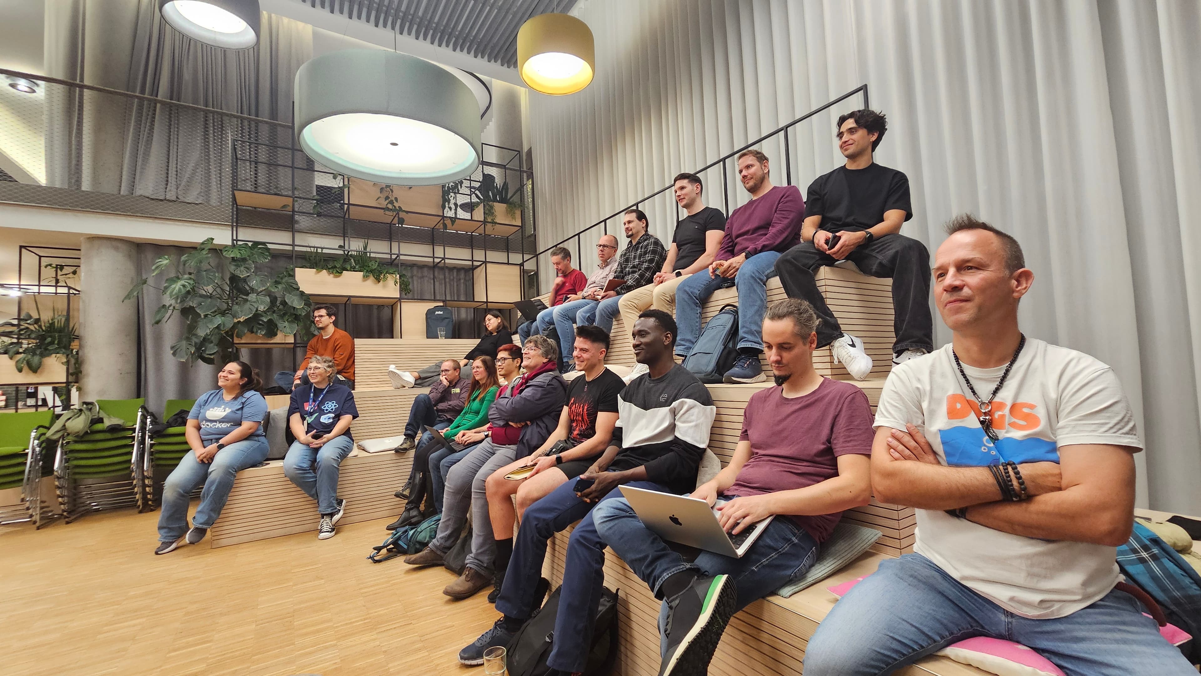 A wide view of attendees watching a presentation from the wooden seating beneath large pendant lights.