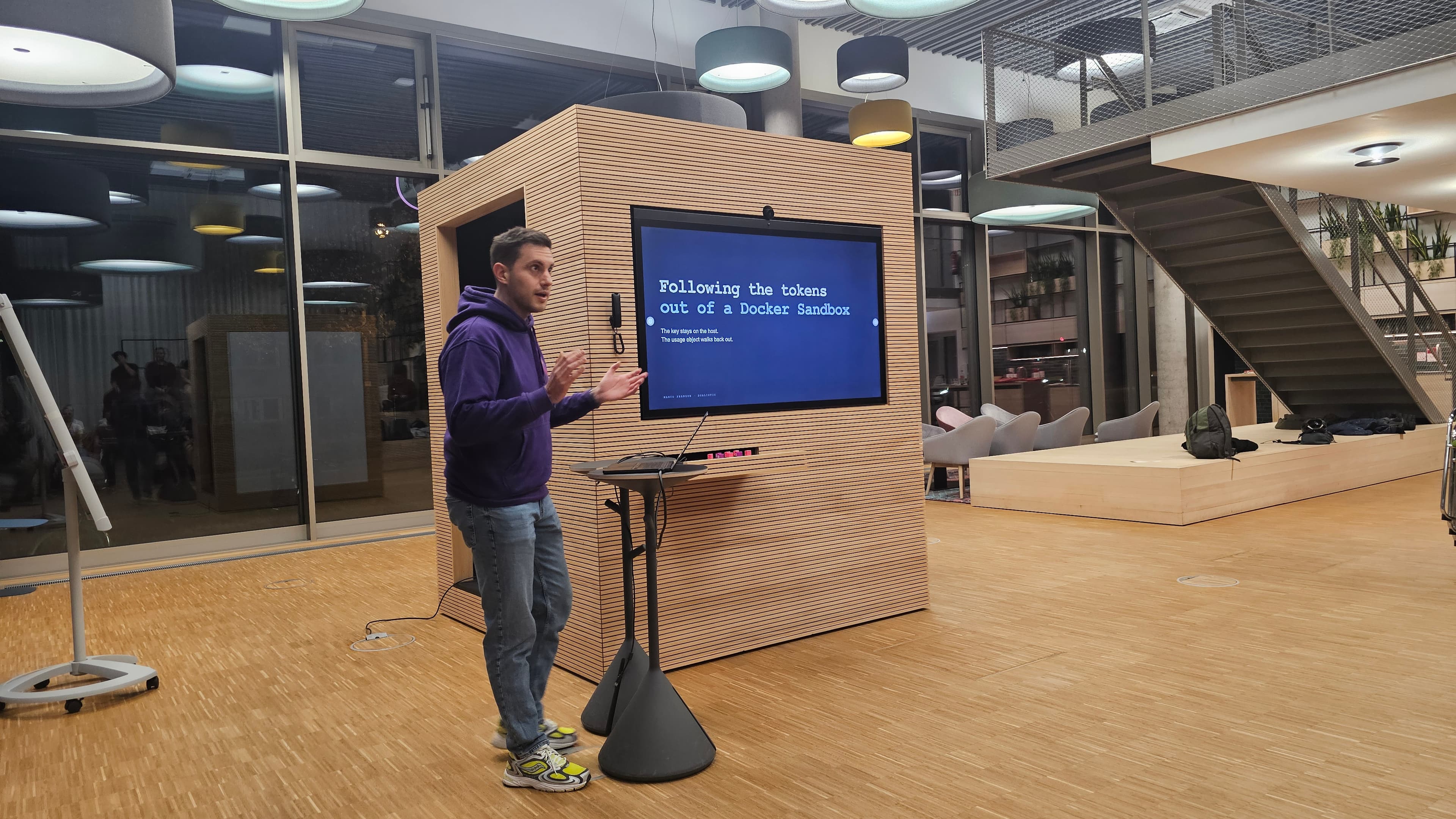 A presenter in a purple hoodie gestures beside a slide titled Following the tokens out of a Docker Sandbox.